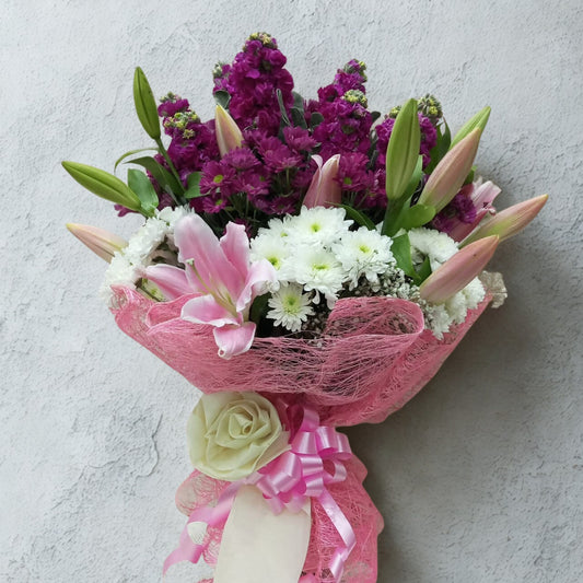 Step into a garden of elegance with this multi-dimensional bouquet featuring layers of purple stalks, playful daisy pops, and standout pink lilies. Each flower speaks a different note in this poetic floral arrangement.