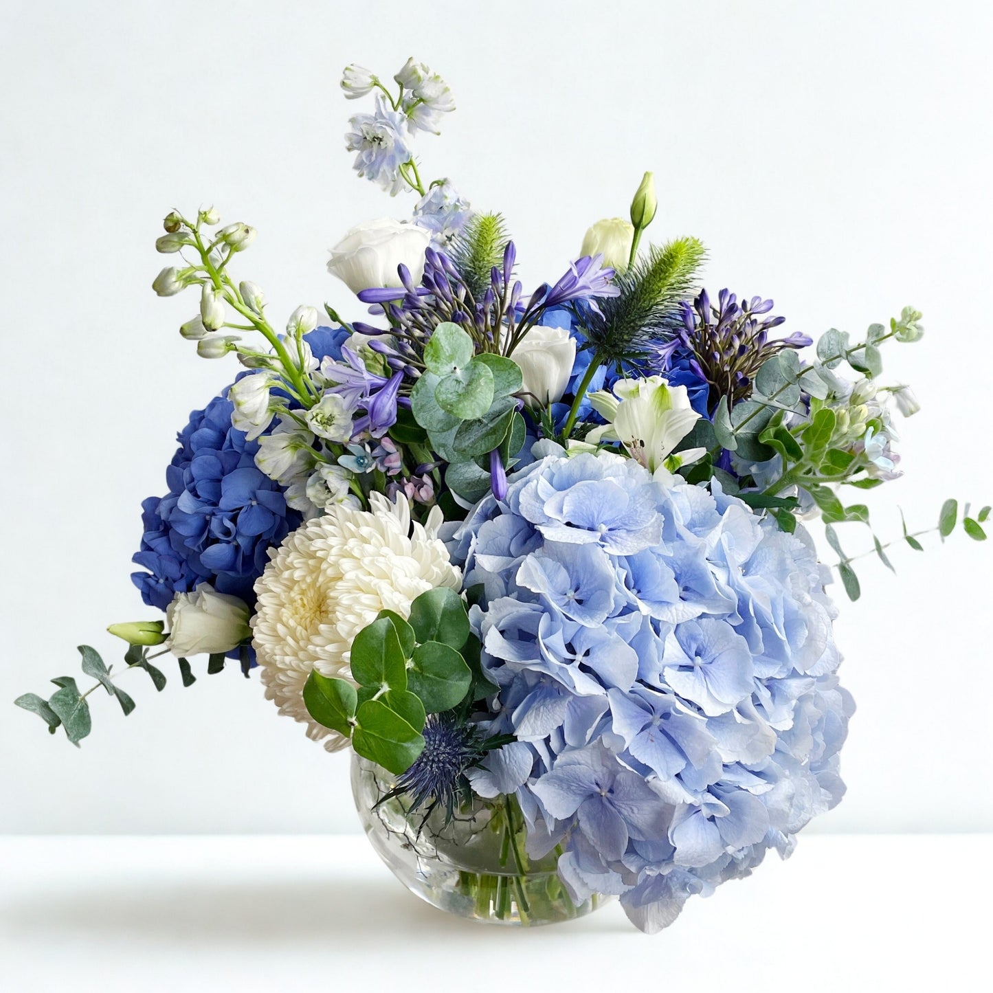 Inspired by the calm of ocean waves and soft clouds, this bouquet features blue and white hydrangeas, carnations, eustoma (lisianthus), orchids, wax flowers, and silver dollar eucalyptus. A serene and elegant arrangement perfect for expressing peace, love, and timeless grace.