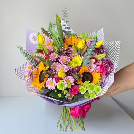 A radiant and cheerful bouquet featuring daisies, sunflowers, lilies, lisianthus (eustoma), and carnations, beautifully accented with green buttons, eucalyptus, and lush green leaves. This vibrant arrangement captures the essence of joy and warmth, perfect for brightening any space or celebrating special moments.