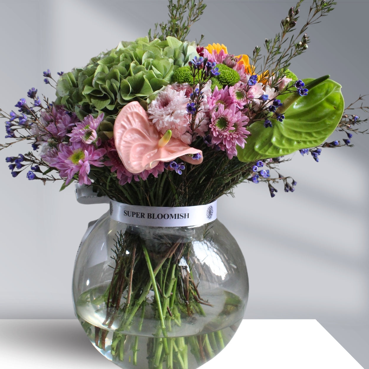 A vibrant floral centerpiece with sunflowers, hydrangeas, pink and white daisies, green buttons, and wax flowers in cheerful hues of yellow, pink, green, and white.