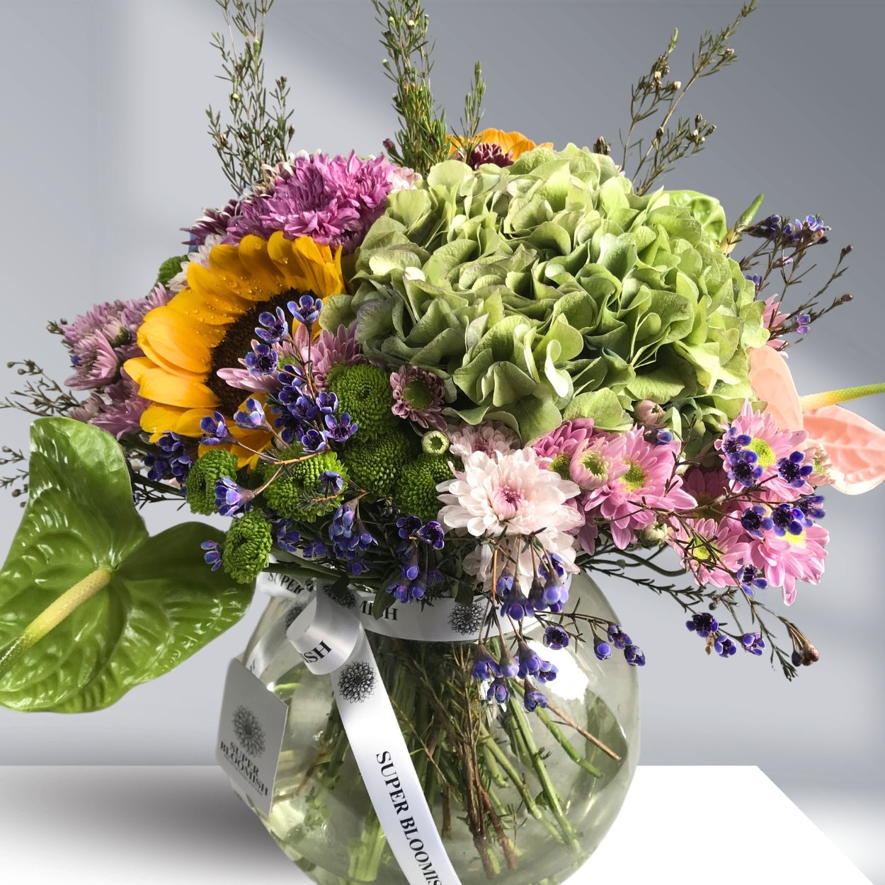 A vibrant floral centerpiece with sunflowers, hydrangeas, pink and white daisies, green buttons, and wax flowers in cheerful hues of yellow, pink, green, and white.