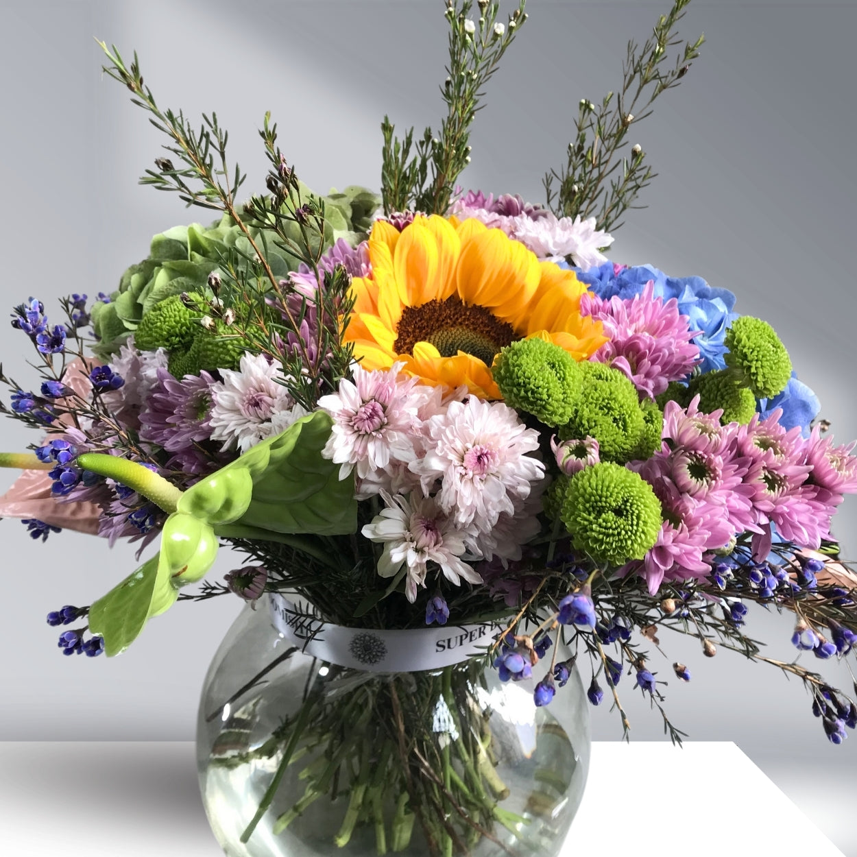 A vibrant floral centerpiece with sunflowers, hydrangeas, pink and white daisies, green buttons, and wax flowers in cheerful hues of yellow, pink, green, and white.