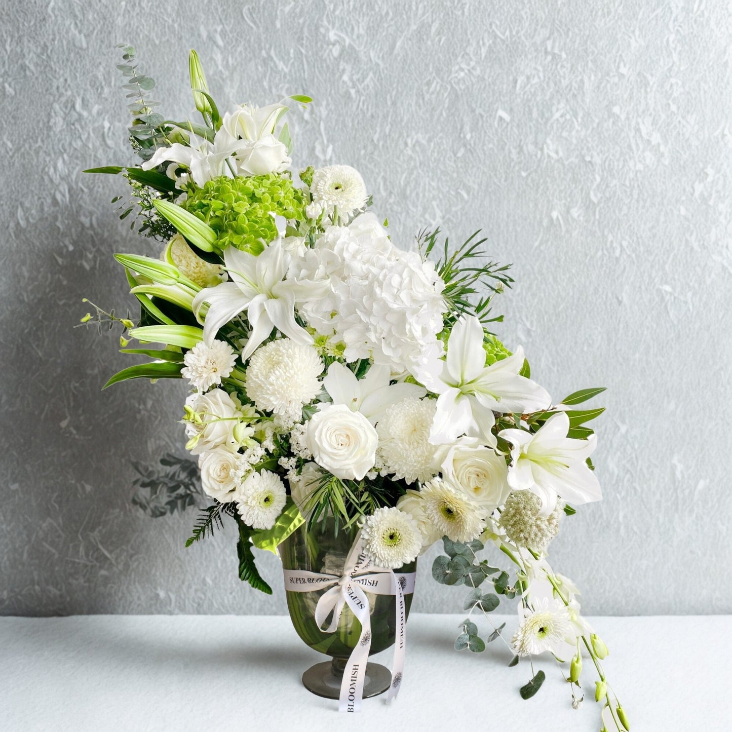 A graceful medley of pure white blooms and lush greenery, this bouquet features hydrangeas, lilies, roses, carnations, daisies, silver dollar eucalyptus, and green fillers. Soft, fresh, and effortlessly elegant—perfect for heartfelt gestures or serene spaces.